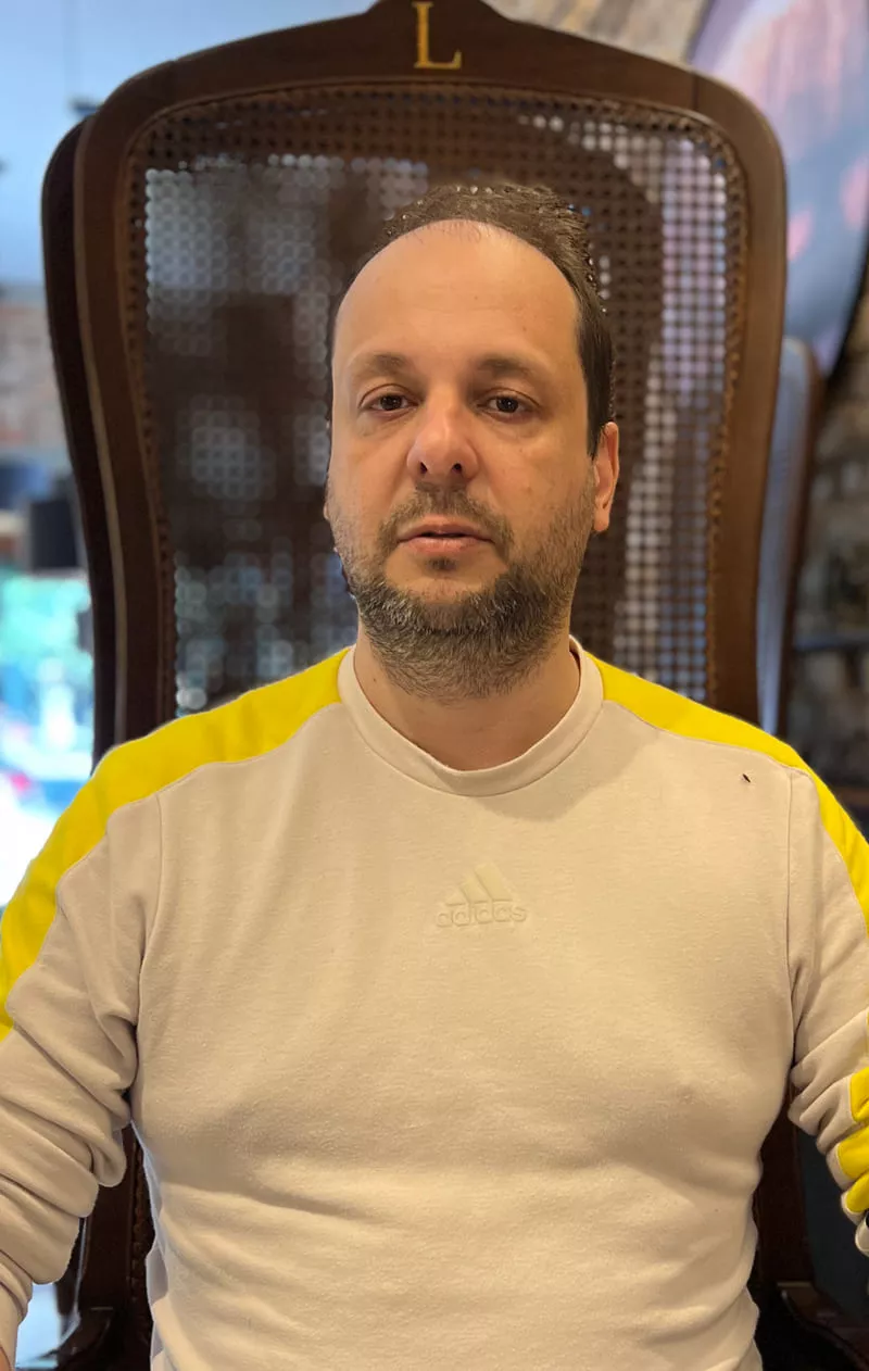 Marcelo Menezes, técnico em refrigeração, de moletom cinza com listras amarelas nos ombros, sentado em cadeira de madeira, olhando para a câmera